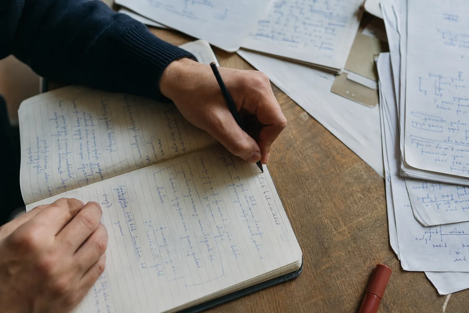 Vue en plongée sur deux mains écrivant au crayon dans un carnet ouvert, feuilles annotées empilées à côté : dans un bilan, la personne travaille elle-même, elle ne se contente pas de remplir un questionnaire.